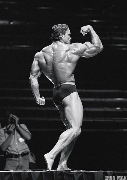Arnold Swarchenegger éppen a pódiumon áll és feszíti a bicepszét és a hátát.