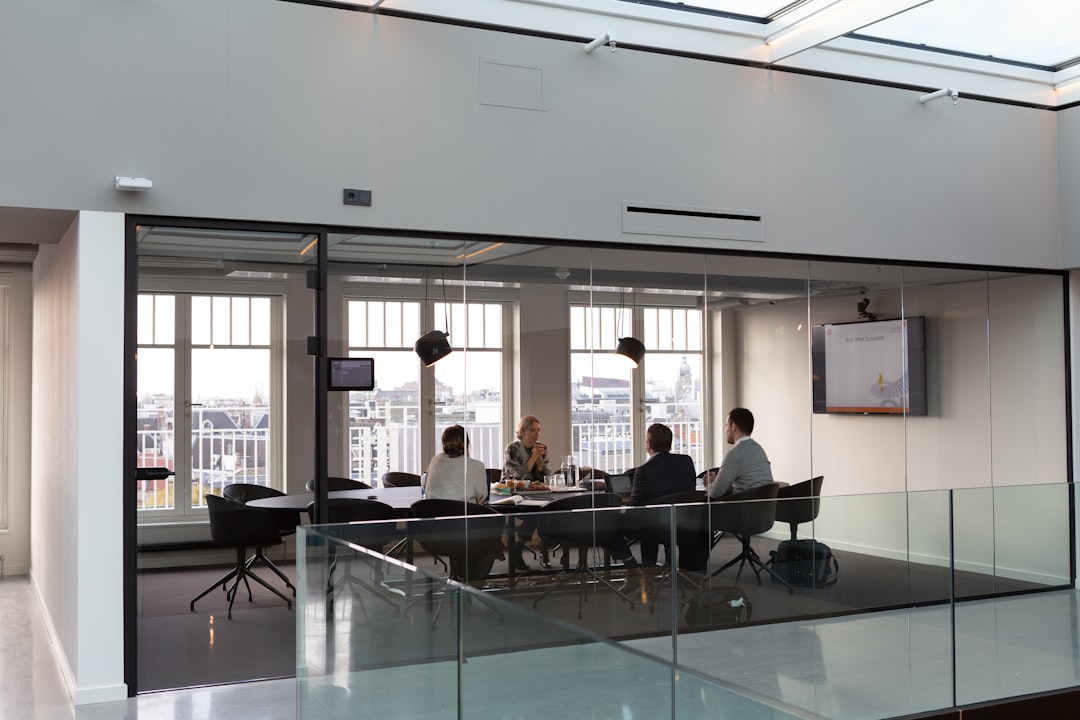 Four people are seated around a conference table in a modern glass-walled meeting room, engaged in discussion with papers, drinks, and a laptop in front of them. The room is bright with natural light from large windows overlooking a cityscape, and a wall-mounted screen displays a presentation slide titled 'Any other business.'