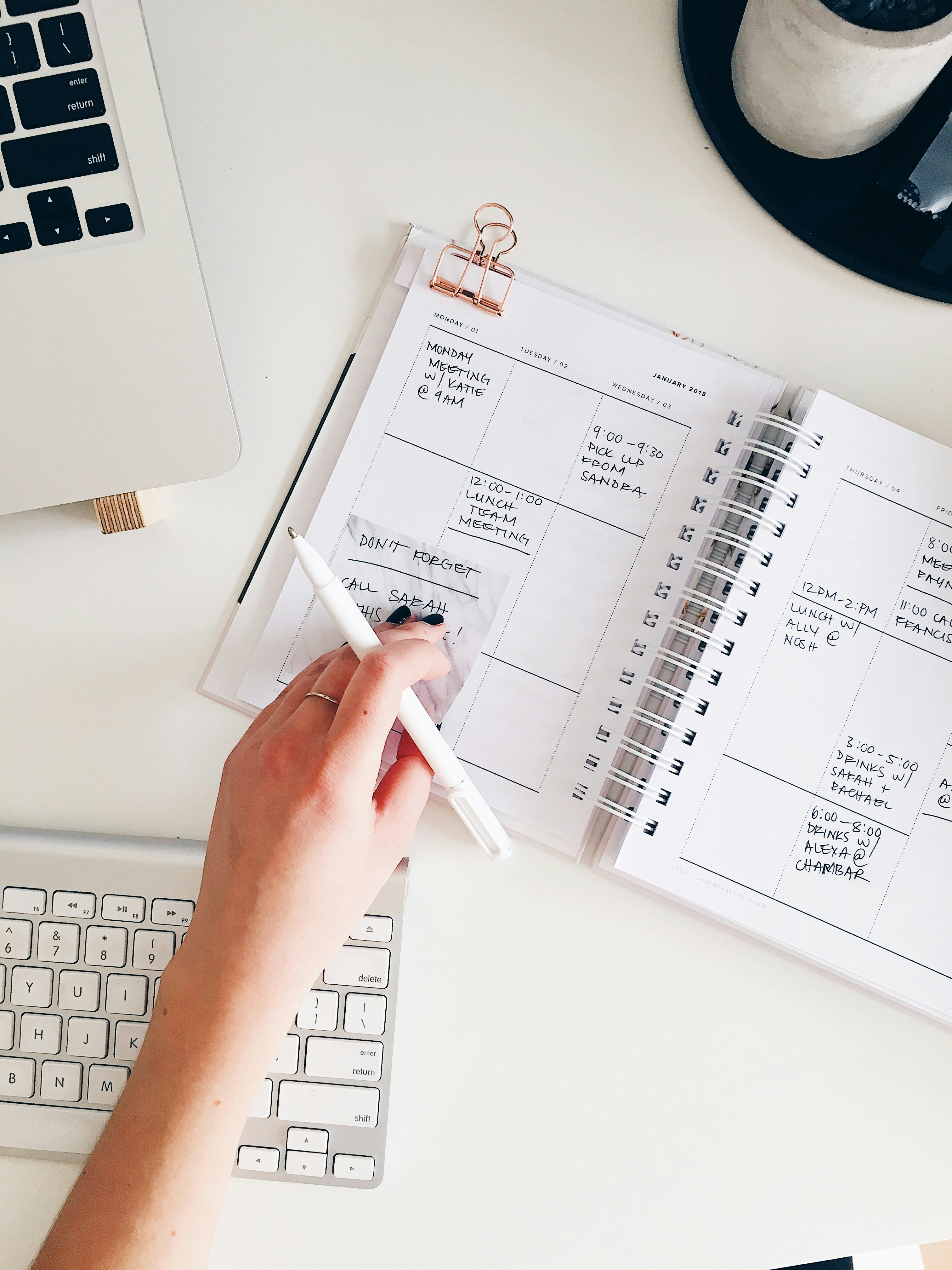 A hand holding a white pen hovers over an open spiral-bound planner on a white desk, showing handwritten notes for the week, with part of a MacBook, an Apple keyboard, and a black tray with a candle and pens nearby. The planner lists events like meetings, a lunch, and drinks with friends, with a rose gold binder clip holding a small note inside.