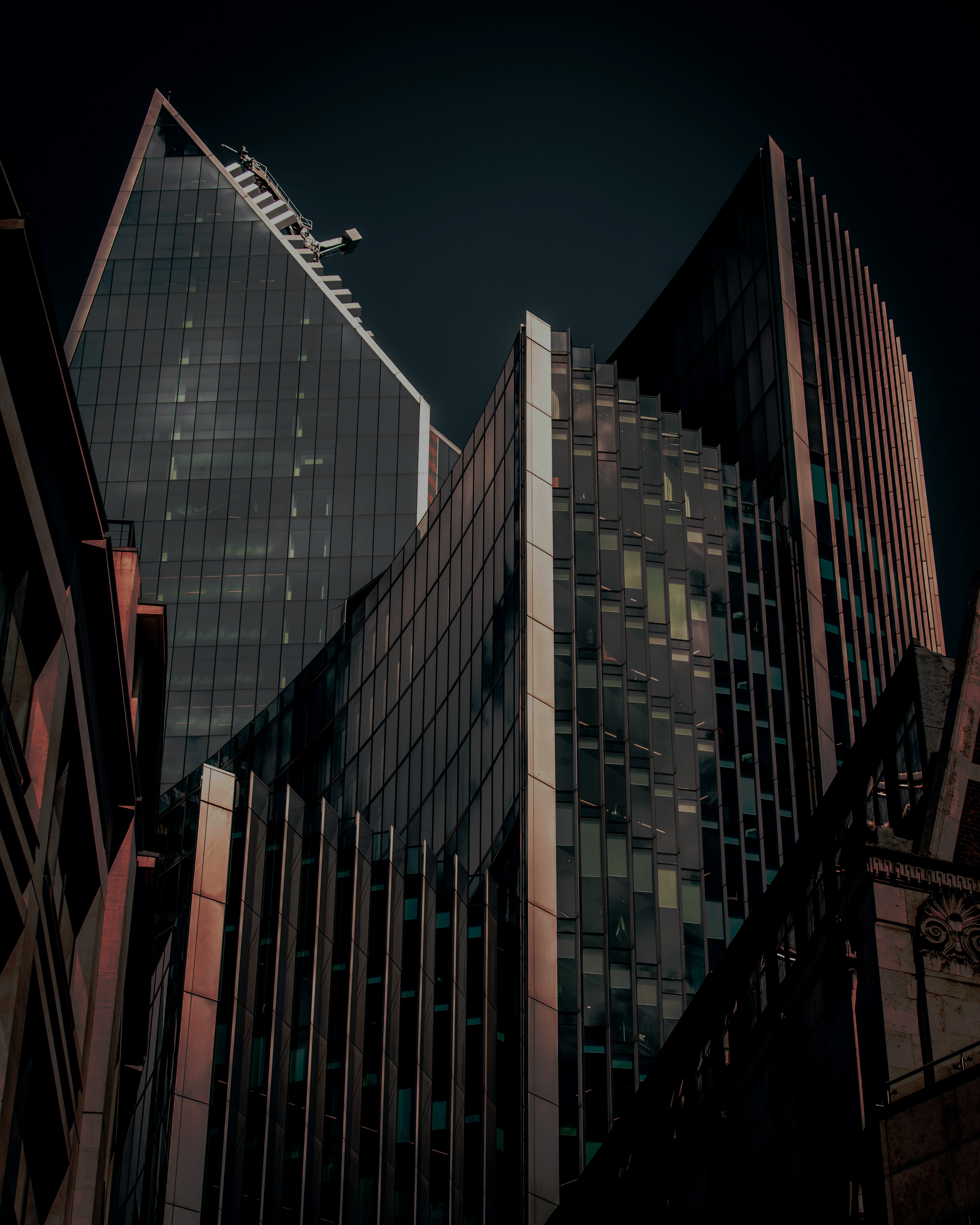 A dramatic view of modern high-rise buildings with sharp, angular glass facades, captured in low light with deep shadows and reflections. The architecture appears sleek and futuristic, with a dark, moody atmosphere against the night sky.