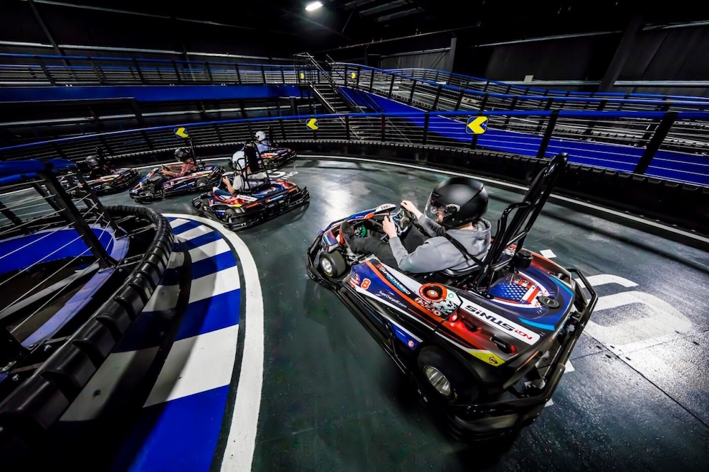 Gokart versenyzők egy éles kanyarban, 5 fő színes sisakokban, biztonsági korlátokkal övezett pályán haladnak különböző versenyautókban