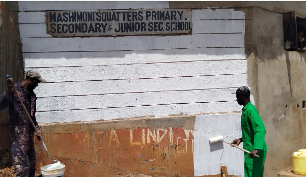 Zöld egyenruhás önkéntes fehérre festi a Mashimoni Squatters Primary & Secondary School falát