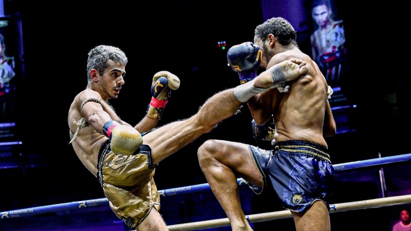 Egy Muay Thai harcos magas rúgással támad ellenfelére a ringben, aki ezt karjával blokkolja.