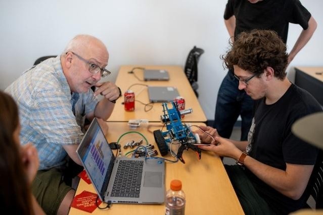 Photo of hobbyists' gathering, 3 people sitting together at a desk, with another standing in the background. One person at the table is assembling an electronic project.